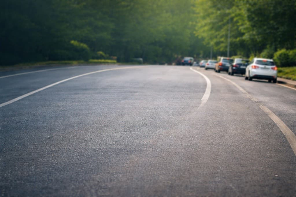 An open road flanked by trees with parked cars in the distance